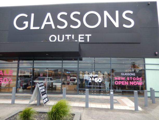 A wide-angle view of a modern Glassons Outlet storefront in a commercial mall, featuring a large black brand sign and bright interior lighting reflecting through large glass panels.